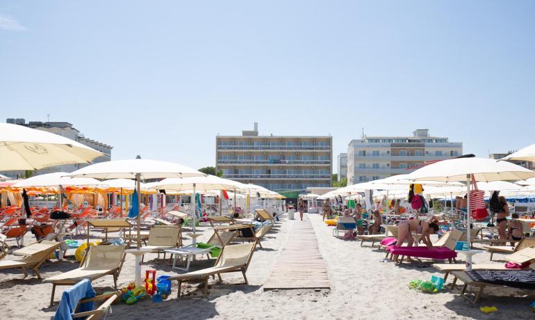 Belebter Strand mit Sonnenschirmen und Liegen, Gebäude im Hintergrund.