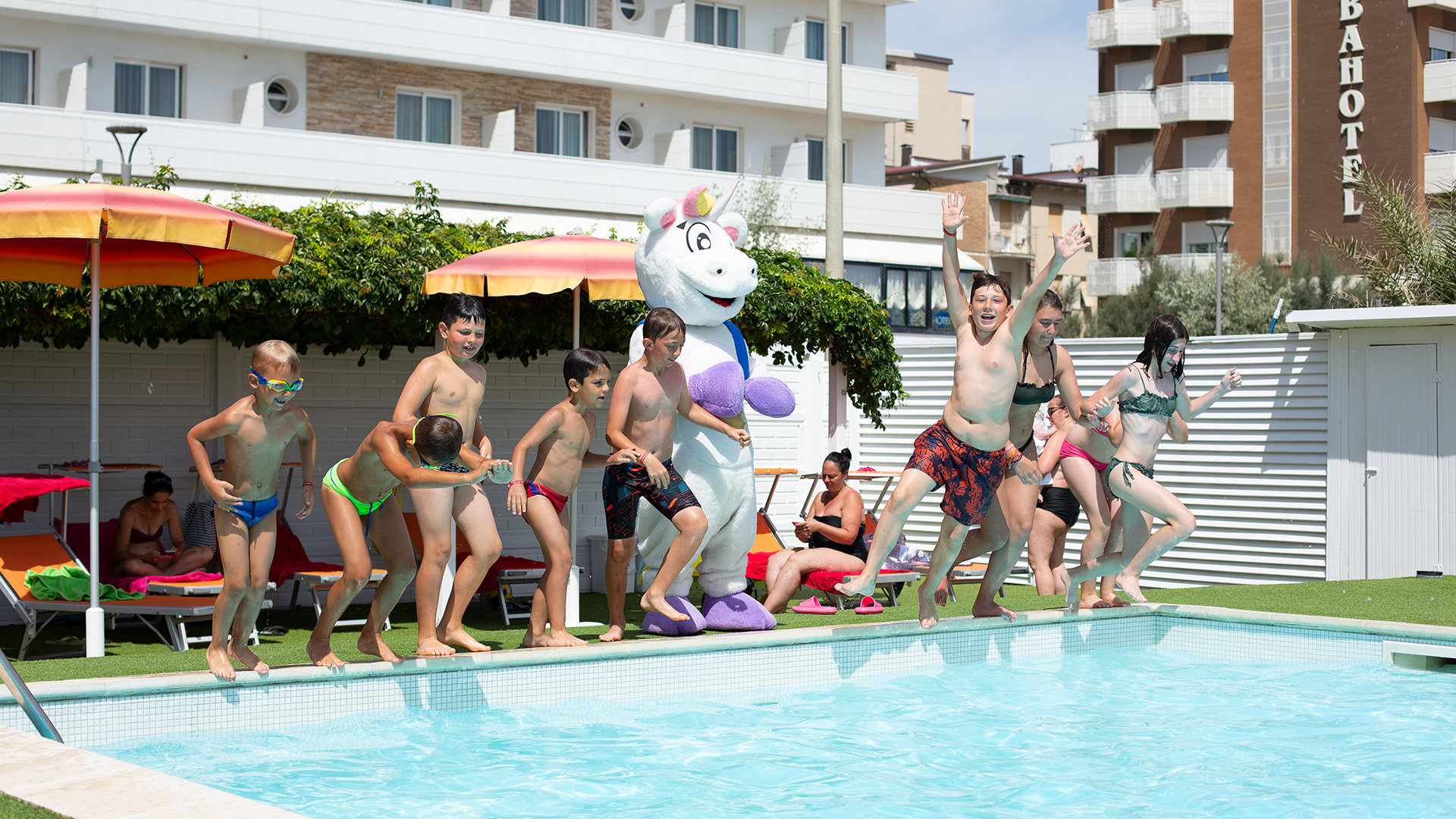 Bambino e uomo in piscina con braccioli, ombrelloni colorati sullo sfondo.