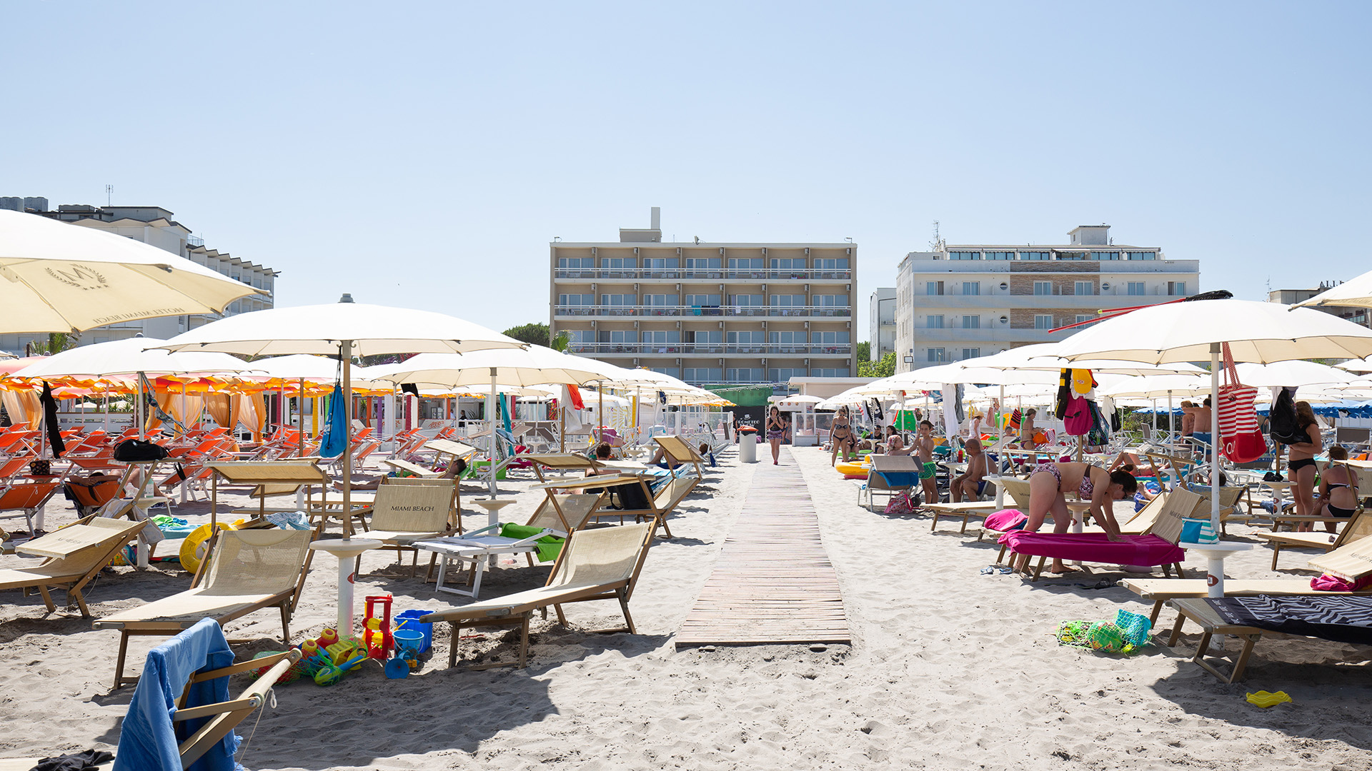 Famiglia cammina sulla spiaggia, tra ombrelloni e sdraio, verso il mare.