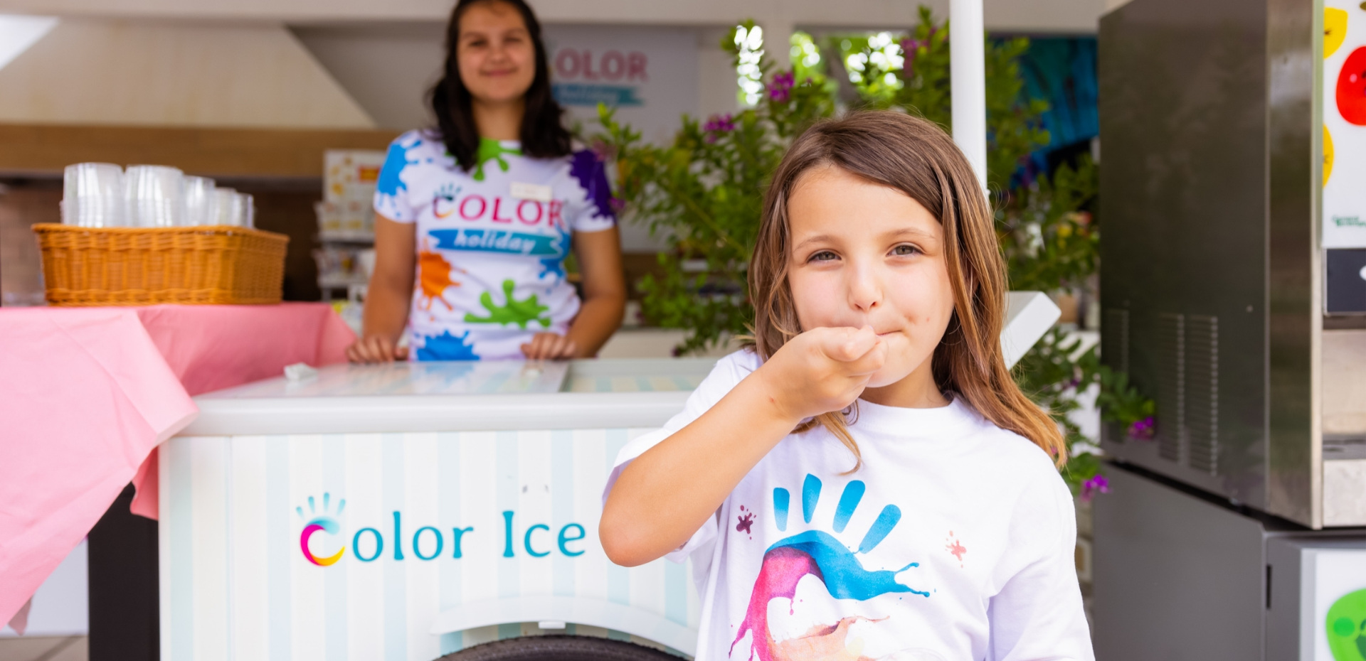 Bambina felice mangia gelato, sorridente venditrice sullo sfondo.