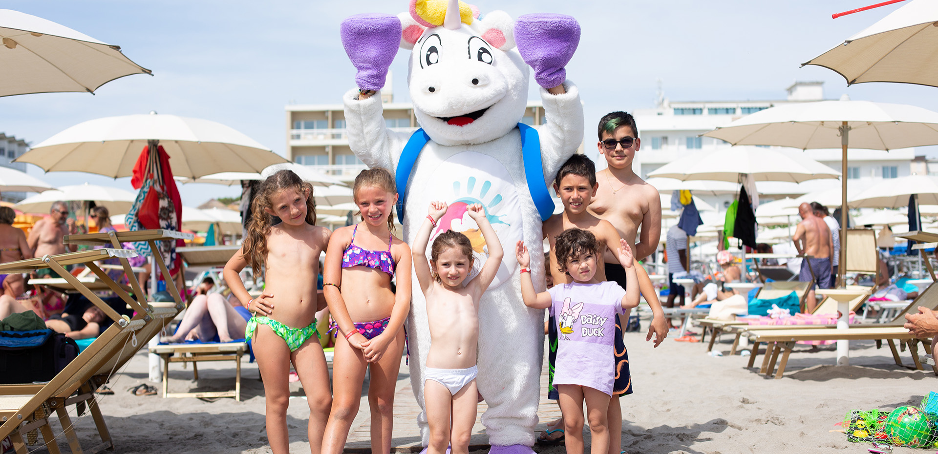 Bambini sorridenti in spiaggia con un costume da unicorno, sotto ombrelloni.